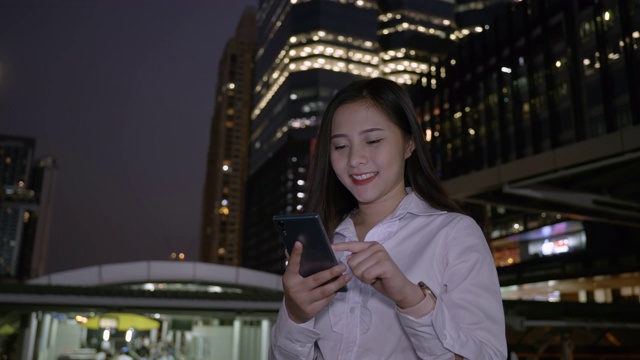 年轻微笑的亚洲商务女性使用智能手机在现代化的市中心在泰国曼谷的夜晚。通过手机进行技术交流视频素材