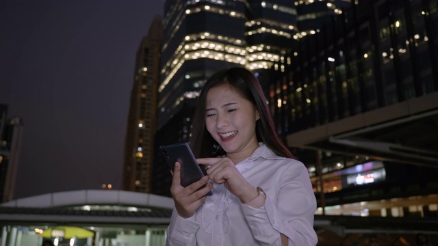 兴奋的商业亚洲妇女庆祝好消息检查智能手机在现代城市中心在泰国曼谷晚上。技术沟通，成功靠手机视频素材