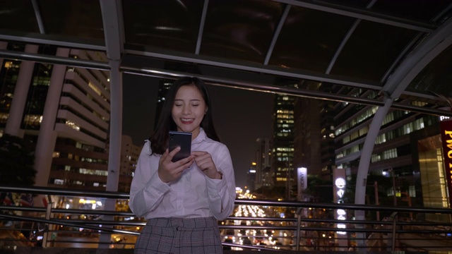 年轻微笑的亚洲商务女性使用智能手机在现代化的市中心在泰国曼谷的夜晚。通过手机进行技术交流视频素材