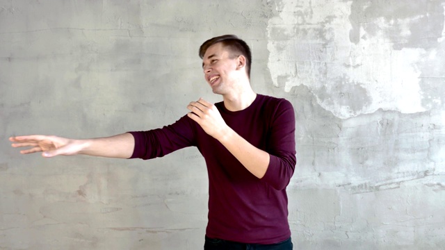 一个身穿紫红色套头衫的年轻人站在一堵灰色的墙上，边笑边挥手。平均计划。视频下载