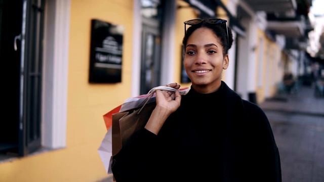 一个迷人的混血女孩的肖像，微笑着走在街上与五颜六色的购物袋。快乐的年轻女子走在购物后的高档街道城市，橱窗购物视频素材