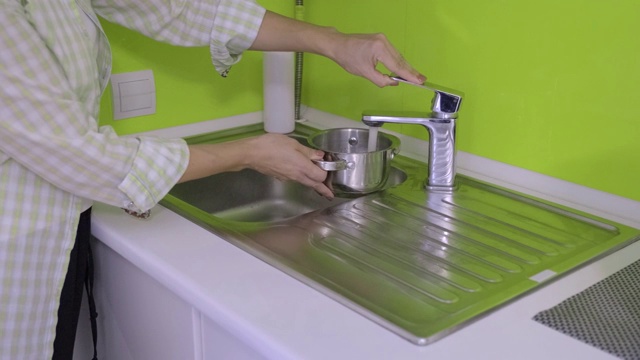 一个女人在浅绿色的厨房里用金属小平底锅倒水，视频下载