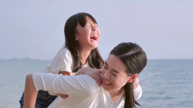快乐的亚洲女婴骑在妈妈的背上，在沙滩上享受海浪带来的乐趣。积极的父母和人在暑假与孩子户外活动。幸福家庭假日，家庭，生活方式，人，人寿保险，假期视频下载