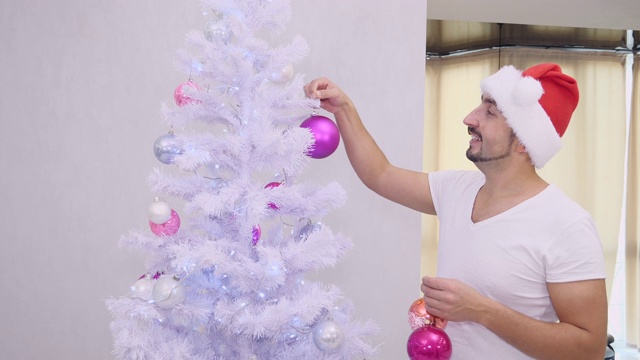 戴着圣诞帽的滑稽大胡子男人正在把圣诞树上的小饰品都拿走。视频下载