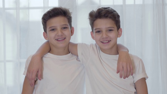 高加索双胞胎看着相机微笑的肖像。一对双胞胎兄弟穿着相似的白色t恤，手搭在彼此的肩膀上聊天。兄弟姐妹在室内摆姿势。视频下载