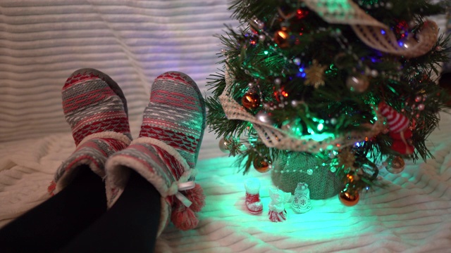 彩色的圣诞树和女孩的腿在温暖的冬天拖鞋在白色的背景。家中温馨圣诞气氛，假日心情概念视频素材
