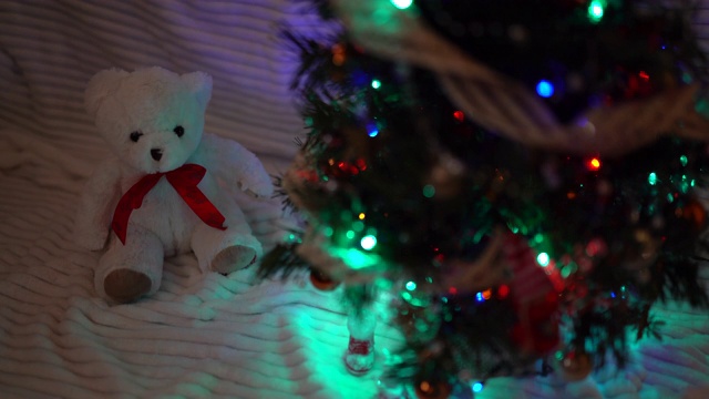黑暗中挂着闪闪发光的花环的小圣诞树。白色小泰迪熊坐在圣诞树下，庆祝的心情和童话的概念视频素材