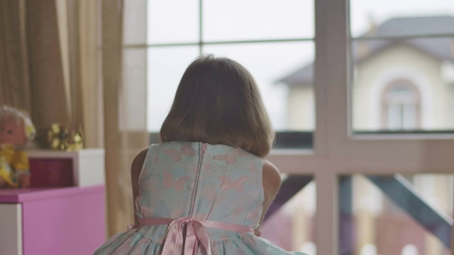 背影特写的白人女孩看着相机的肩膀，签名和转身回窗口。在她的房间里，穿着蝴蝶的孩子坐在粉红色的床上。视频素材