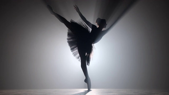 职业芭蕾舞演员在聚光灯下跳芭蕾舞，大舞台上烟雾缭绕。漂亮的年轻女孩穿着黑色芭蕾舞裙在泛光灯的背景。黑色和白色。4 k视频素材