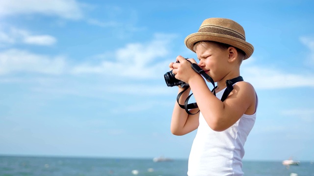 快乐旅行的小男孩戴着帽子在蓝天背景下用相机拍摄海景视频下载