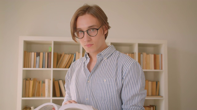 一个年轻成功的白人男学生的特写照片，戴着眼镜，看着相机在图书馆里看书，背景是书架视频素材