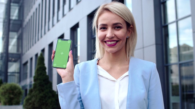 照片中，这位美丽开朗的年轻白种金发女商人站在户外，手里拿着智能手机，然后微笑着向镜头展示一款绿色屏幕的手机。色度键。跟踪运动。视频素材