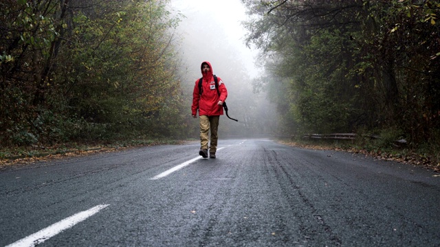 穿着红色夹克和背包的年轻人走在空旷的，废弃的道路上的森林视频下载