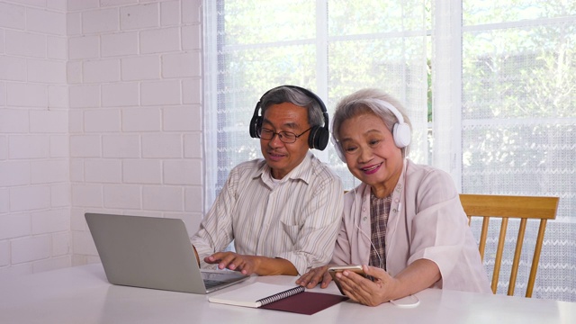 幸福的亚洲老年夫妇使用笔记本电脑和手机，同时享受在家里听音乐，家庭放松和花时间在一起，退休人士和生活方式的概念视频素材