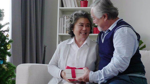 一名年长的亚裔男子在客厅里给恋人送了一个白色礼盒。欢度圣诞，恭贺新年，笑口常开。是退休人员的家庭活动。老年社会的概念视频素材