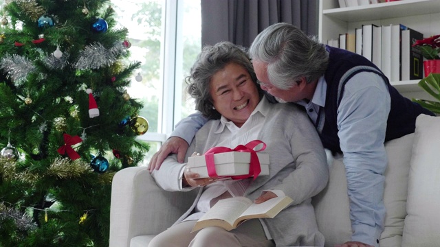 一名年长的亚裔男子在客厅里给恋人送了一个白色礼盒。欢度圣诞，恭贺新年，笑口常开。是退休人员的家庭活动。老年社会的概念视频素材
