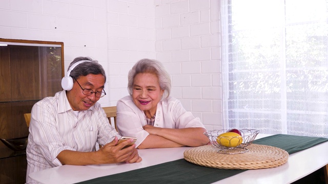 幸福的亚洲老年夫妇使用手机和享受听音乐，而坐在家里，家庭放松和花时间在一起，退休人士和生活方式的概念视频素材