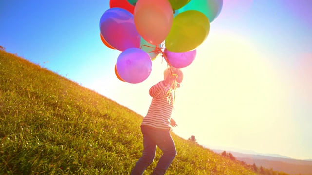一个小男孩拿着一堆气球，和他们一起跑过阳光下的草地视频素材
