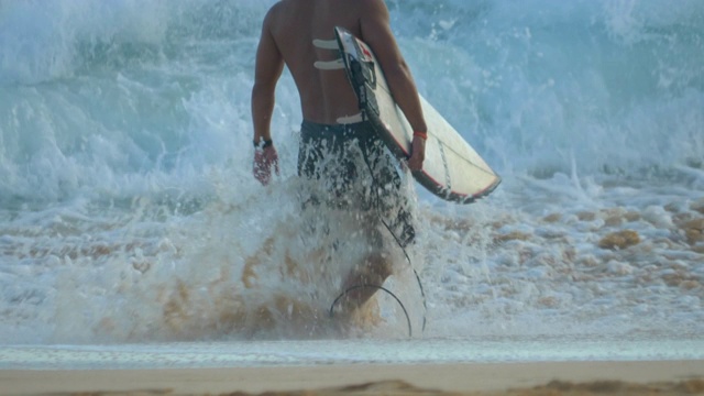 一个冲浪者带着他的冲浪板在夏威夷瓦胡岛的沙滩上冲浪。——慢动作视频素材