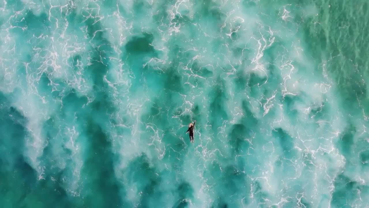 一名冲浪者在清澈的绿色海水中划桨和冲浪海浪的无人机视图。——慢动作视频下载