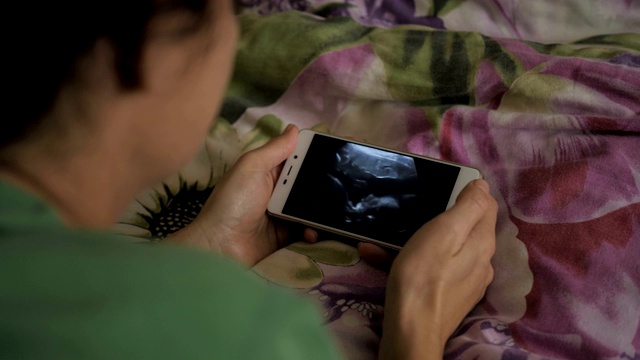 孕妇躺在床上用智能手机看超声波扫描。怀孕晚期妊娠。孕妇在手机上看超声波视频。妇科分娩诞生。视频素材
