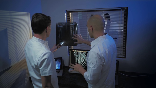 在控制室，医生和放射科医生一边看程序一边讨论诊断，在后台病人正在接受MRI或CT扫描程序。视频素材