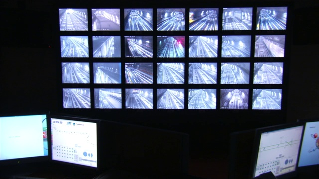 在两台电脑上方的墙上安装了多个安全屏幕，每台电脑有两台显示器。视频下载