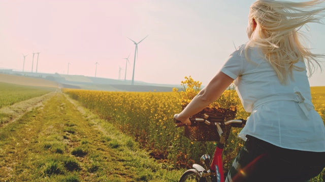 一名女子骑着自行车沿着油菜籽田行驶，远处有风力涡轮机视频素材