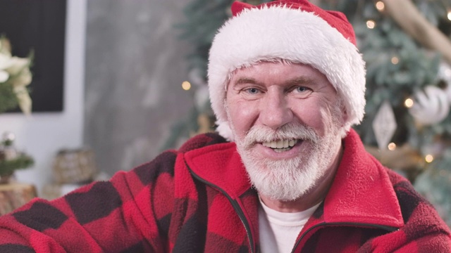 特写的老白人男子在红色圣诞帽看着照相机和眨眼。积极的圣诞老人摆姿势的背景装饰新年树。电影4k ProRes总部。视频下载