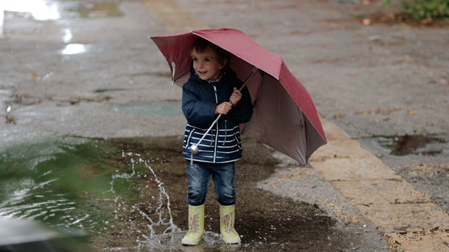 一个男孩打着伞穿着胶靴站在雨中的水坑里。视频素材