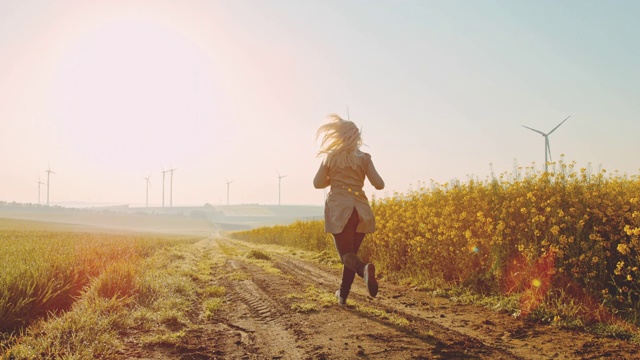 沿着油菜籽田跑步的妇女，远处有风力涡轮机视频素材