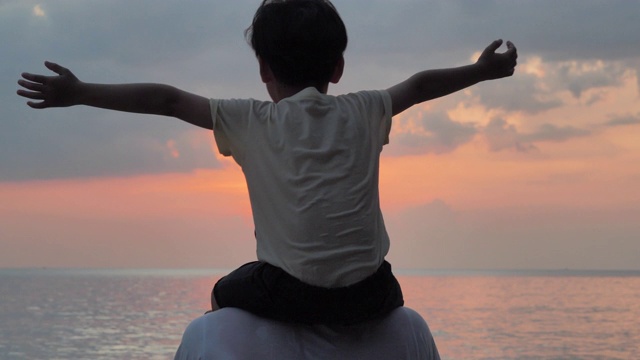 亚洲男婴骑在父亲脖子上的剪影。在一个夏天的日落时分，爸爸和小男孩一起在户外玩耍。一家人在海滩上看日落。假日旅行的概念。假期视频下载