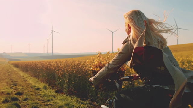 女人骑着自行车沿着油菜籽田走，远处有风力发电机视频素材