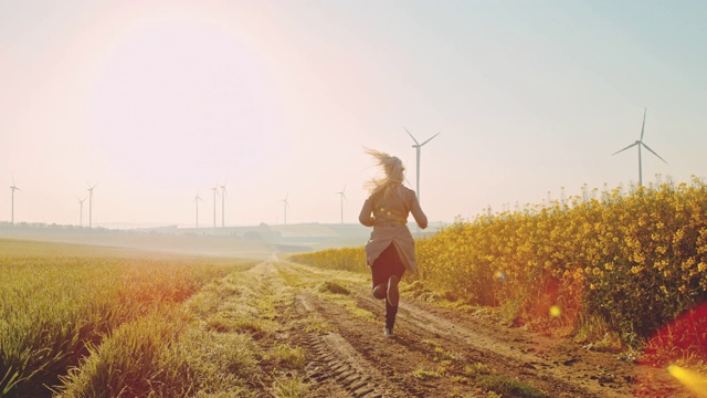 超级慢MO -时间扭曲效应妇女跑在油菜田与风力涡轮机在远处视频素材