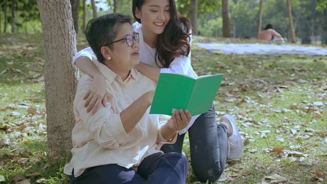 亚洲家庭一位母亲正在公园的树下看书。一个十几岁的女儿来拥抱爱，微笑，友谊，同居和关心老年人的健康是必要的。视频素材