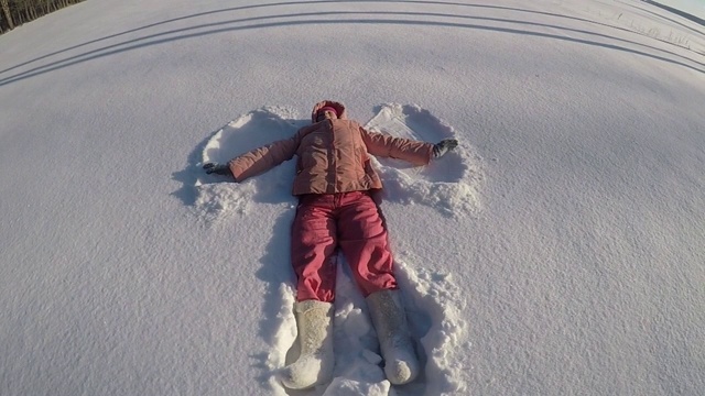 一个穿着桃色夹克和粉色裤子的女人躺在雪堆里，做了一个雪天使。冷淡的晴朗的日子。慢动作视频素材