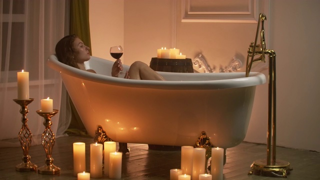女人躺在泡泡浴中，用蜡烛的光放松和喝葡萄酒。浪漫放松的芳香疗法氛围。浴室用蜡烛视频素材