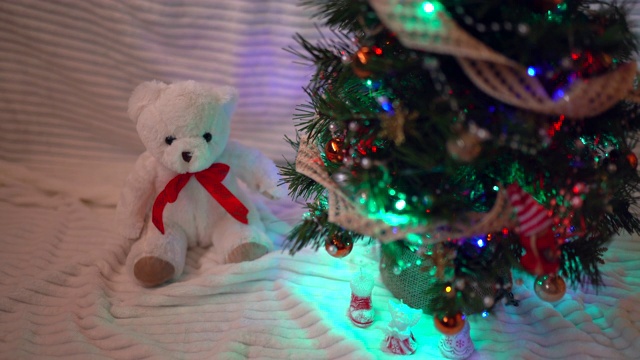 在黑暗中，圣诞树上挂着五颜六色的球和玩具。白色小泰迪熊坐在圣诞树下，庆祝的心情和童话的概念视频素材