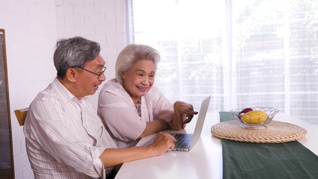 幸福的亚洲老年夫妇使用笔记本电脑在家里，家庭放松和花时间在一起在家里，退休人士和生活方式的概念视频素材