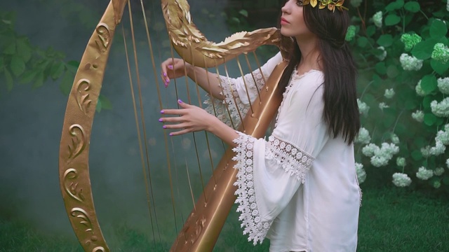美丽的精灵女孩弹奏着古老的金色竖琴。公主在春天开花的花园里。视频素材