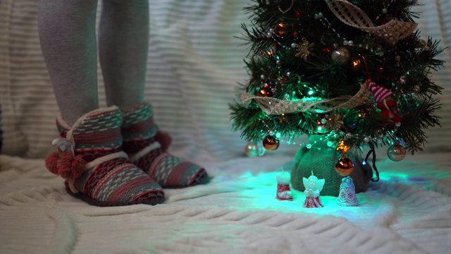 令人惊叹的装饰圣诞树和苗条女孩的腿在温暖的冬天拖鞋呆在树下。魔法照亮晚会前的圣诞节。圣诞树上闪闪发光的花环视频素材