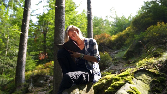 一个年轻女子坐在森林里的岩石上看书视频素材