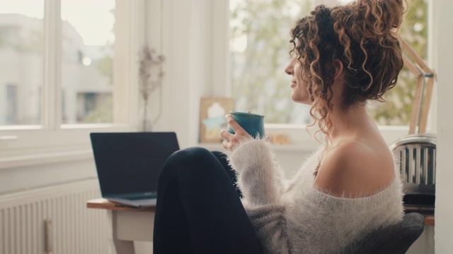 年轻女子坐在家里的笔记本电脑前喝茶视频素材
