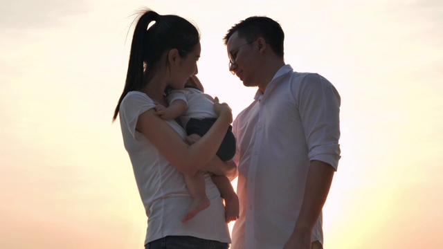 剪影亚洲母亲抱着婴儿的儿子和父亲亲吻他的甜蜜沿着沙滩上的日落海浪边缘。视频素材