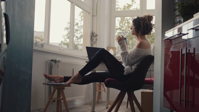 年轻女子坐在家里的笔记本电脑前喝咖啡视频下载