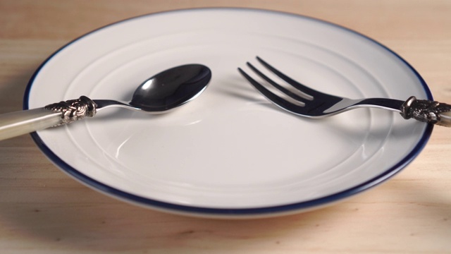 木桌上有一个边缘有蓝色条纹的白色盘子，配有甜点匙和叉子视频下载