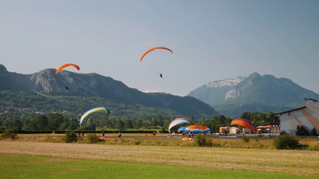 橙色滑翔伞在阿尔卑斯山脉的着陆跑道上降落。男性和女性乘坐滑翔伞在田野上空飞行视频素材