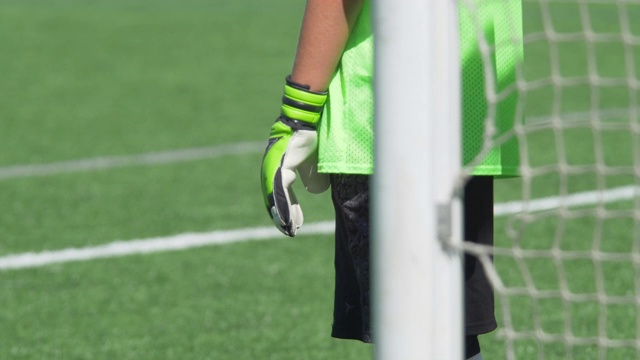 一个戴着绿色手套的男孩在草坪上踢少年足球守门员。——慢动作视频下载