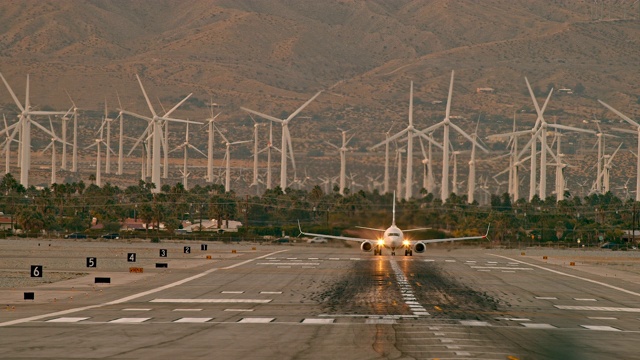 LS客机从机场跑道起飞，空气中充满了喷气发动机的热烟雾，背景是风力发电场的旋转涡轮机视频下载