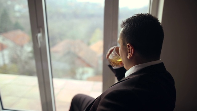 年轻的衣着讲究的商人放松地坐在窗边，一边看风景，一边喝着威士忌视频下载